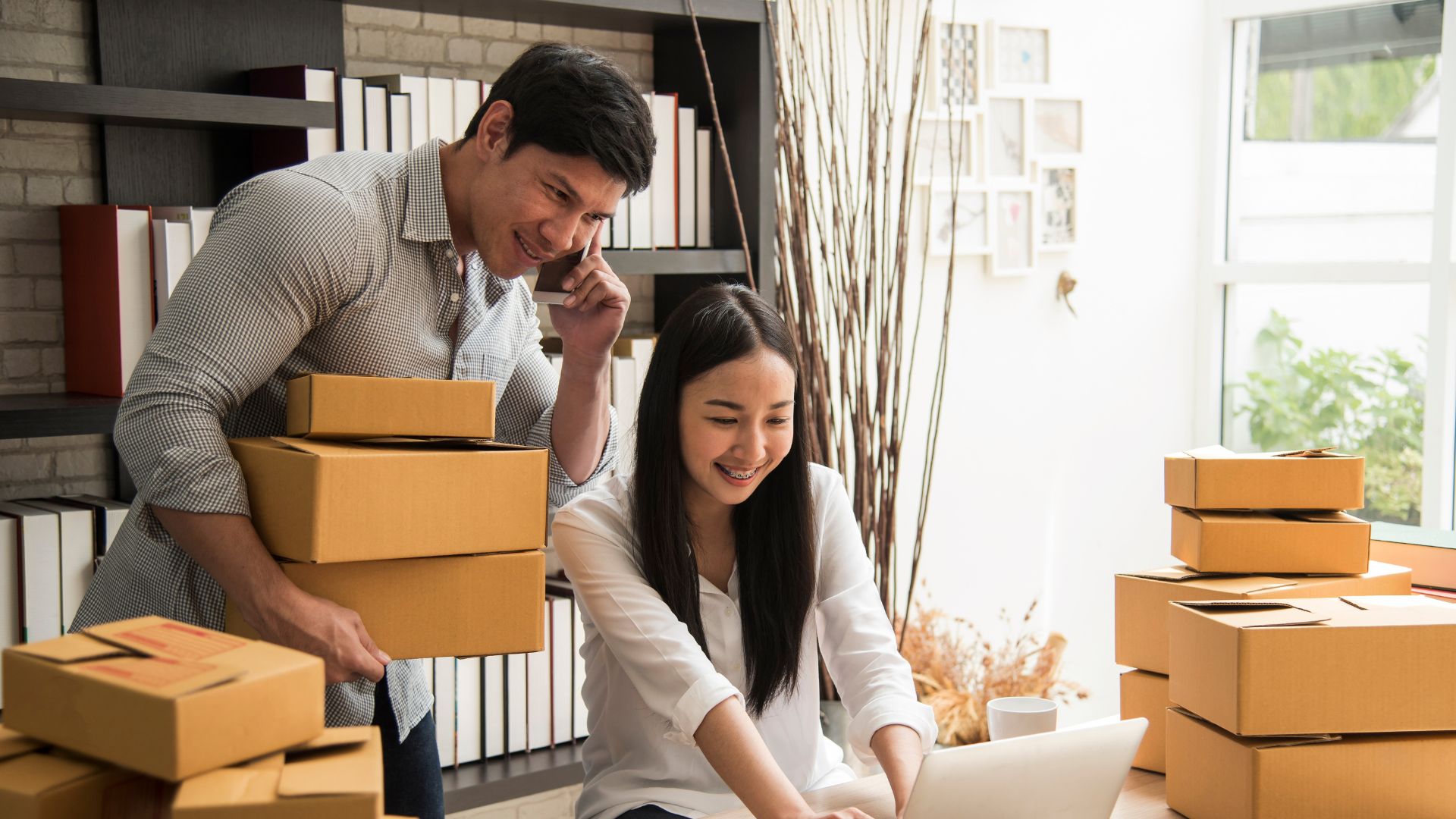 A man and a woman surrounded by boxes, the man is on the phone the woman on a laptop